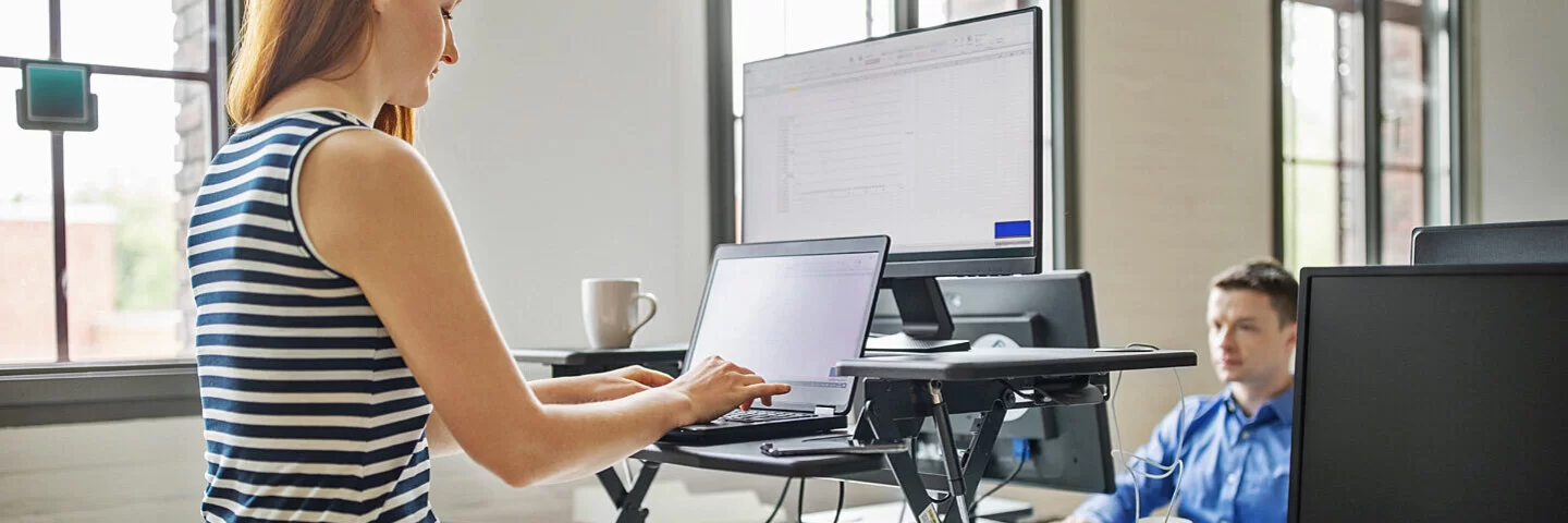 Frau steht an ihrem ergonomischen Stehschreibtisch im Büro. Sie arbeitet an ihrem Computer im Stehen.