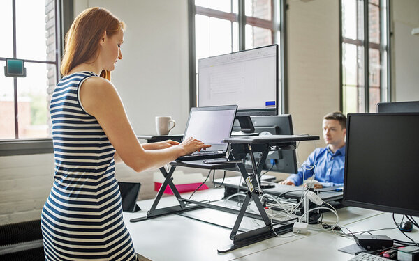 Frau steht an ihrem ergonomischen Stehschreibtisch im Büro. Sie arbeitet an ihrem Computer im Stehen.