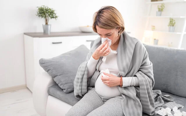 Eine schwangere Frau sitzt auf einem Sofa und putzt sich die Nase.