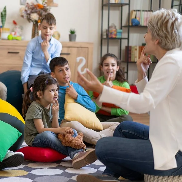 Kinder sitzen auf dem Boden oder auf Sitzsäcken und legen ihren Zeigefinger auf die Unterlippe. Mit einer Therapeutin üben sie die richtige Aussprache.