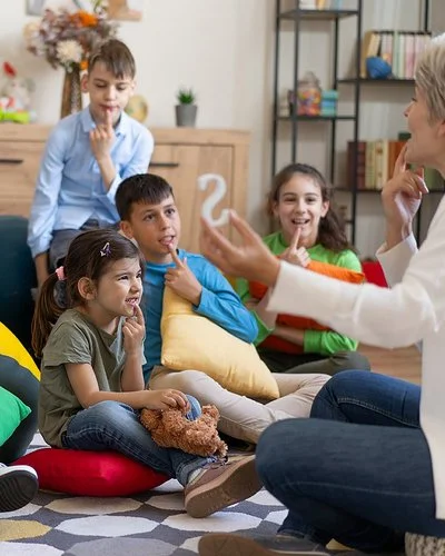 Kinder sitzen auf dem Boden oder auf Sitzsäcken und legen ihren Zeigefinger auf die Unterlippe. Mit einer Therapeutin üben sie die richtige Aussprache.