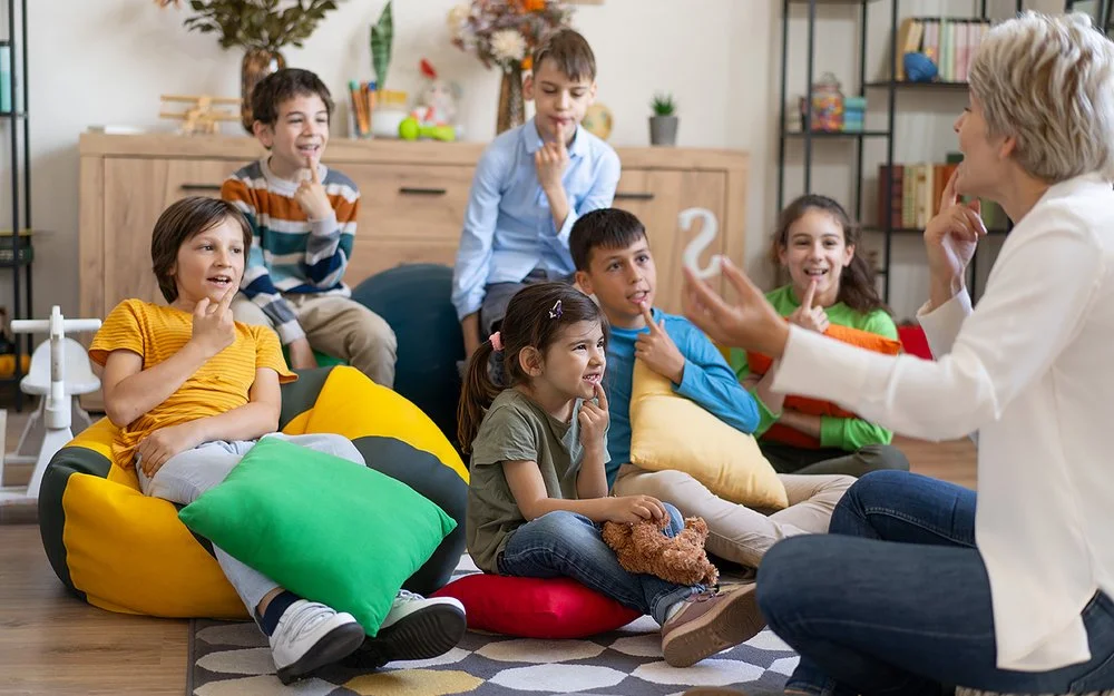Kinder sitzen auf dem Boden oder auf Sitzsäcken und legen ihren Zeigefinger auf die Unterlippe. Mit einer Therapeutin üben sie die richtige Aussprache.