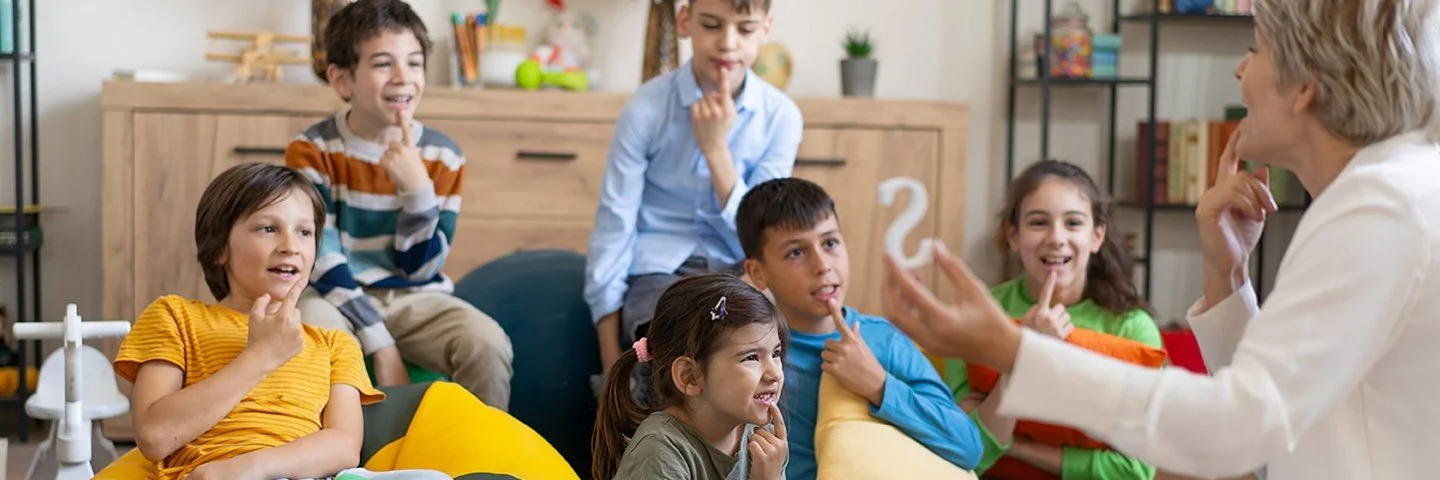 Kinder sitzen auf dem Boden oder auf Sitzsäcken und legen ihren Zeigefinger auf die Unterlippe. Mit einer Therapeutin üben sie die richtige Aussprache.