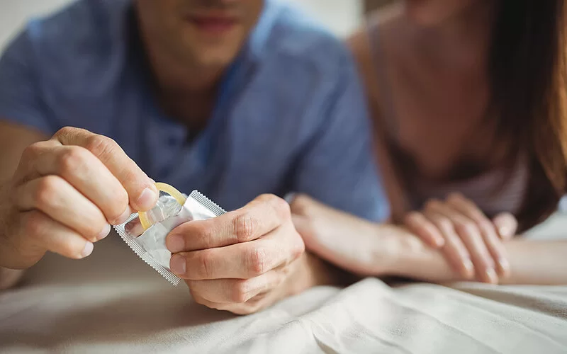 Ein Mann und eine Frau liegen im Bett. Der Mann nimmt ein Kondom aus seiner Verpackung.