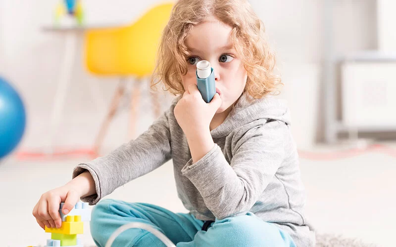 Kleines Mädchen mit blonden Locken sitzt auf dem Boden und inhaliert Kortison mit ihrem Asthmaspray.