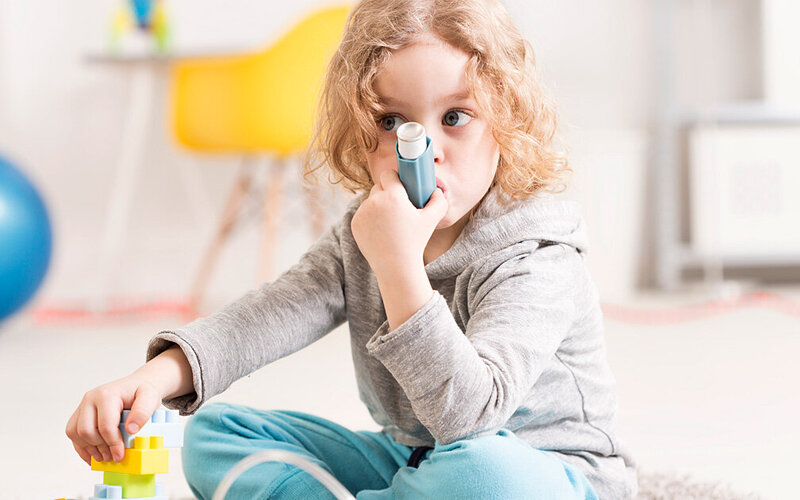 Kleines Mädchen mit blonden Locken sitzt auf dem Boden und inhaliert Kortison mit ihrem Asthmaspray.