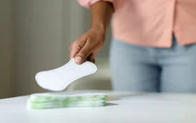 Großaufnahme einer jungen Frau, die mit der rechten Hand von einem Stapel auf einem Tisch eine Slipeinlage greift.