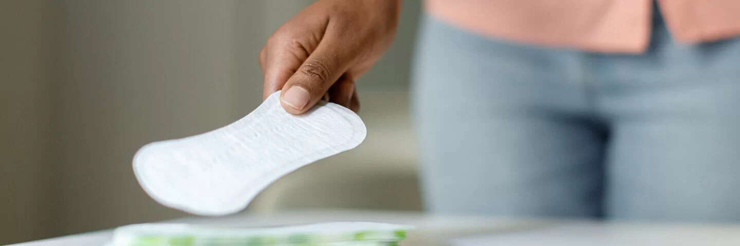 Großaufnahme einer jungen Frau, die mit der rechten Hand von einem Stapel auf einem Tisch eine Slipeinlage greift.