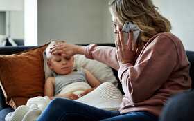 Ein Junge ist an einer Kinderkrankheit erkrankt und liegt auf dem Sofa. Die Mutter fühlt seine Stirn und telefoniert.