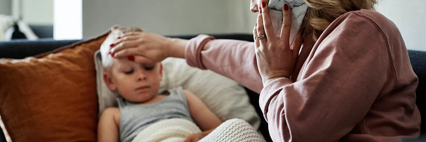 Ein Junge ist an einer Kinderkrankheit erkrankt und liegt auf dem Sofa. Die Mutter fühlt seine Stirn und telefoniert.