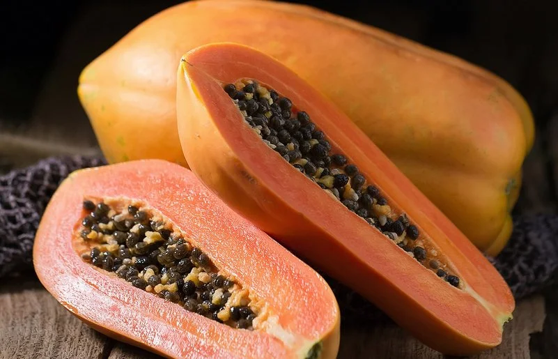 Eine ganze und eine aufgeschnittene Papaya liegen auf einem Holzbrett.