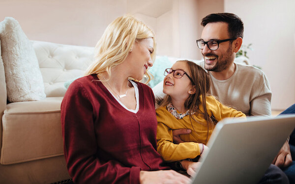Eine Familie sitzt mit einem Laptop auf dem Schoß der Mutter auf dem Boden, Tochter und Vater tragen eine Brille.