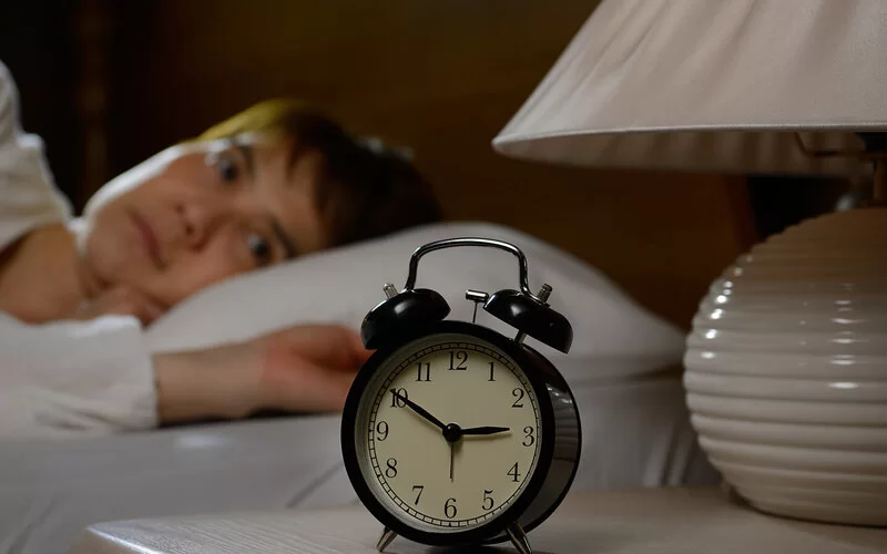 Eine Frau findet keinen Schlaf und liegt mit offenen Augen im Bett. Ein Wecker auf dem Nachttisch zeigt 2.50 Uhr an.