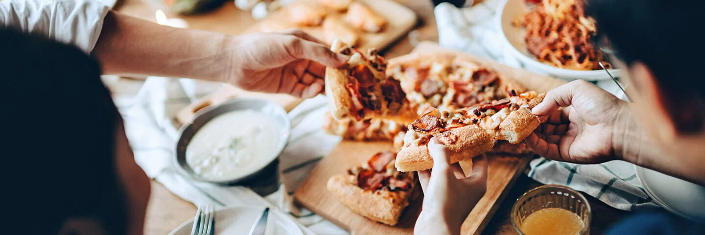 Eine Gruppe junger Menschen isst von einem vollgedeckten Tisch Pizza, Teigtaschen mit Dips und Salat.