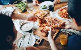 Eine Gruppe junger Menschen isst von einem vollgedeckten Tisch Pizza, Teigtaschen mit Dips und Salat.