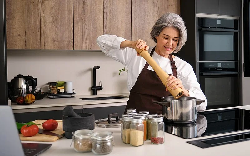 Eine Frau mittleren Alters steht an einem Herd und mahlt Pfeffer aus einer großen Pfeffermühle in einen Topf. Neben der Herdplatte stehen weitere Gewürzgläser, im Hintergrund ist eine Küchenzeile zu erkennen.