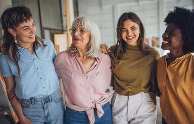 Vier lachende Frauen verschiedenen Alters und Herkunft, die frontal abgebildet sind und sich unterhaken.