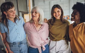 Vier lachende Frauen verschiedenen Alters und Herkunft, die frontal abgebildet sind und sich unterhaken.