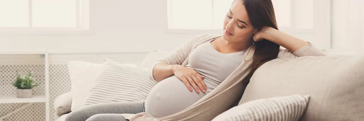 Eine junge, schwangere Frau sitzt auf dem Sofa und streichelt sich über den Bauch.