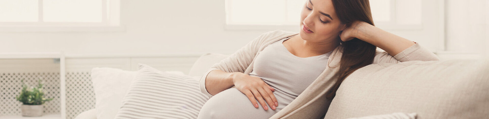 Eine junge, schwangere Frau sitzt auf dem Sofa und streichelt sich über den Bauch.