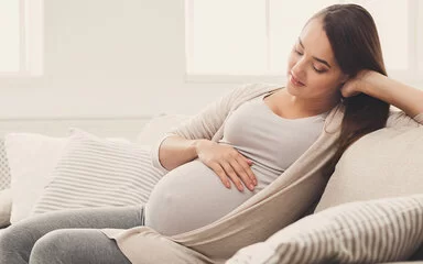 Eine junge, schwangere Frau sitzt auf dem Sofa und streichelt sich über den Bauch.