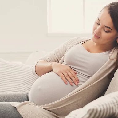 Eine junge, schwangere Frau sitzt auf dem Sofa und streichelt sich über den Bauch.