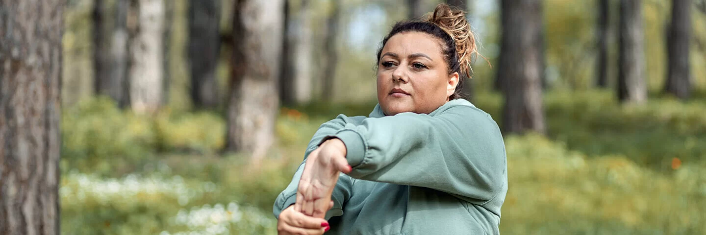 Eine mehrgewichtige Frau dehnt sich in einer Joggingpause im Wald.