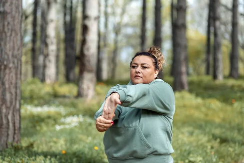 Eine mehrgewichtige Frau dehnt sich in einer Joggingpause im Wald.