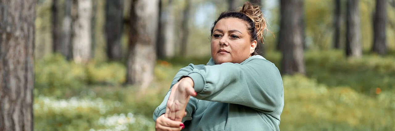 Eine mehrgewichtige Frau dehnt sich in einer Joggingpause im Wald.