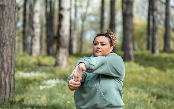 Eine mehrgewichtige Frau dehnt sich in einer Joggingpause im Wald.