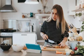 Eine junge Frau steht in der Küche mit einem Messer in der Hand und schaut auf den Bildschirm eines Tablets.