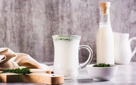 Ein Glas mit Ayran steht auf einem Tisch. Daneben liegt ein Holzbrett mit frischen Kräutern und einem Tuch. Auf dem Tisch stehen noch ein kleines Schälchen mit Kräutern, ein Krug und eine Flasche mit Ayran.