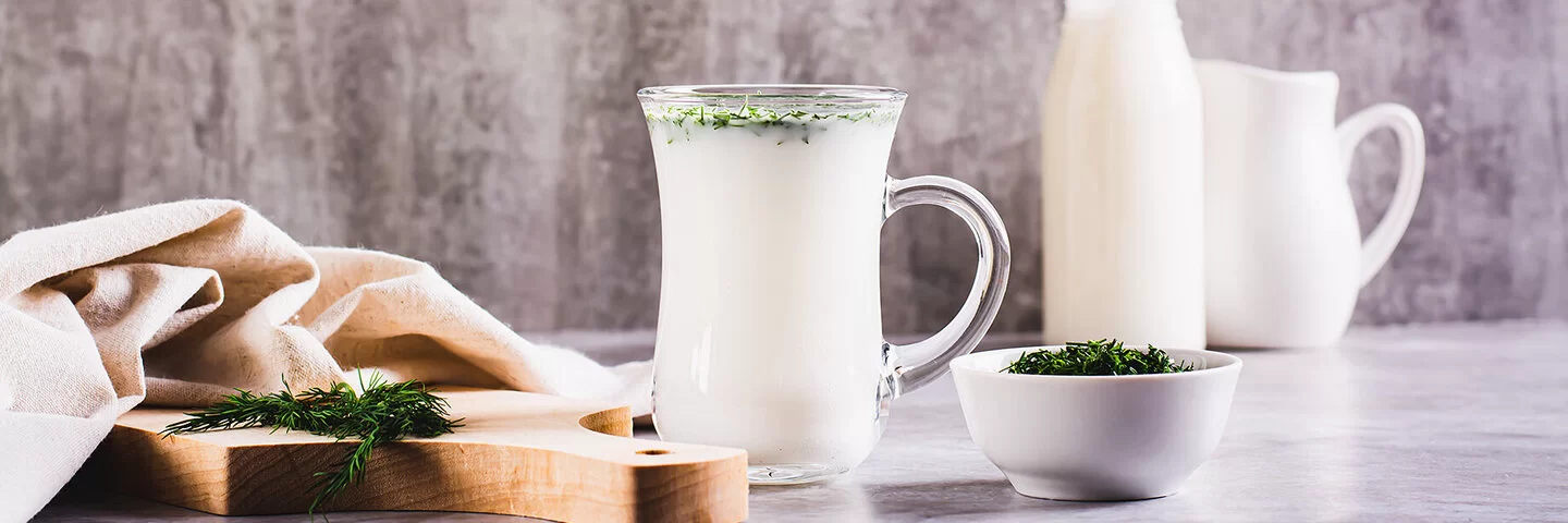 Ein Glas mit Ayran steht auf einem Tisch. Daneben liegt ein Holzbrett mit frischen Kräutern und einem Tuch. Auf dem Tisch stehen noch ein kleines Schälchen mit Kräutern, ein Krug und eine Flasche mit Ayran.