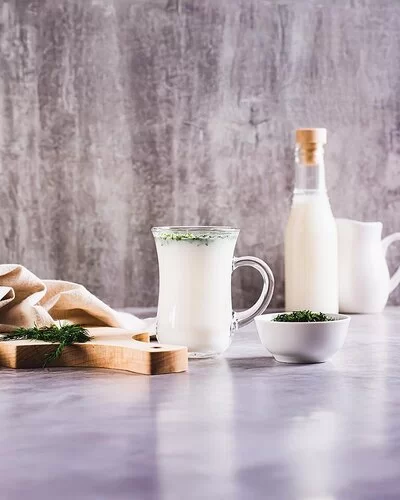 Ein Glas mit Ayran steht auf einem Tisch. Daneben liegt ein Holzbrett mit frischen Kräutern und einem Tuch. Auf dem Tisch stehen noch ein kleines Schälchen mit Kräutern, ein Krug und eine Flasche mit Ayran.