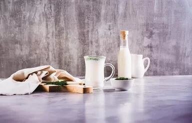 Ein Glas mit Ayran steht auf einem Tisch. Daneben liegt ein Holzbrett mit frischen Kräutern und einem Tuch. Auf dem Tisch stehen noch ein kleines Schälchen mit Kräutern, ein Krug und eine Flasche mit Ayran.