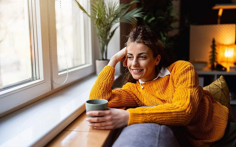 Eine Frau schaut mit einer Tasse in der Hand entspannt aus dem Fenster.