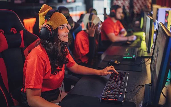 Eine junge Frau sitzt mit Kopfhörern an einem PC. Hinter ihr erkennt man weitere junge Menschen vor Computern. Sie alle tragen die gleichen, orangenen T-Shirts.