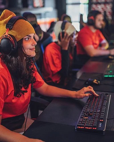 Eine junge Frau sitzt mit Kopfhörern an einem PC. Hinter ihr erkennt man weitere junge Menschen vor Computern. Sie alle tragen die gleichen, orangenen T-Shirts.