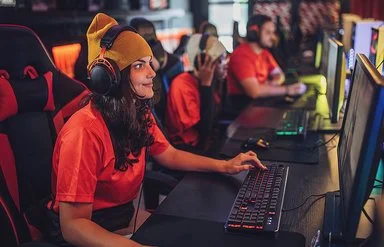 Eine junge Frau sitzt mit Kopfhörern an einem PC. Hinter ihr erkennt man weitere junge Menschen vor Computern. Sie alle tragen die gleichen, orangenen T-Shirts.