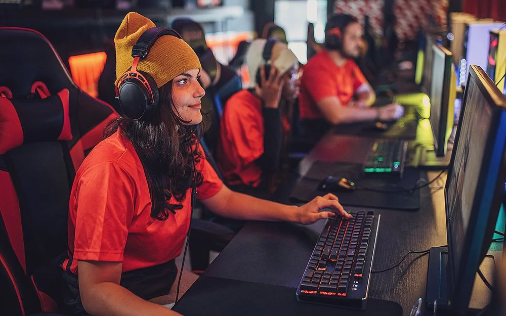 Eine junge Frau sitzt mit Kopfhörern an einem PC. Hinter ihr erkennt man weitere junge Menschen vor Computern. Sie alle tragen die gleichen, orangenen T-Shirts.