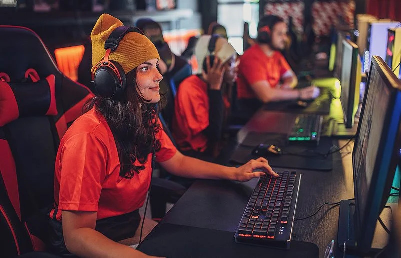 Eine junge Frau sitzt mit Kopfhörern an einem PC. Hinter ihr erkennt man weitere junge Menschen vor Computern. Sie alle tragen die gleichen, orangenen T-Shirts.