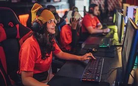 Eine junge Frau sitzt mit Kopfhörern an einem PC. Hinter ihr erkennt man weitere junge Menschen vor Computern. Sie alle tragen die gleichen, orangenen T-Shirts.