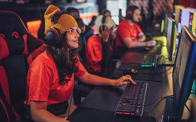 Eine junge Frau sitzt mit Kopfhörern an einem PC. Hinter ihr erkennt man weitere junge Menschen vor Computern. Sie alle tragen die gleichen, orangenen T-Shirts.