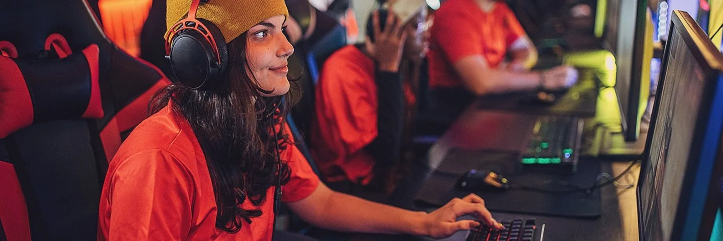 Eine junge Frau sitzt mit Kopfhörern an einem PC. Hinter ihr erkennt man weitere junge Menschen vor Computern. Sie alle tragen die gleichen, orangenen T-Shirts.