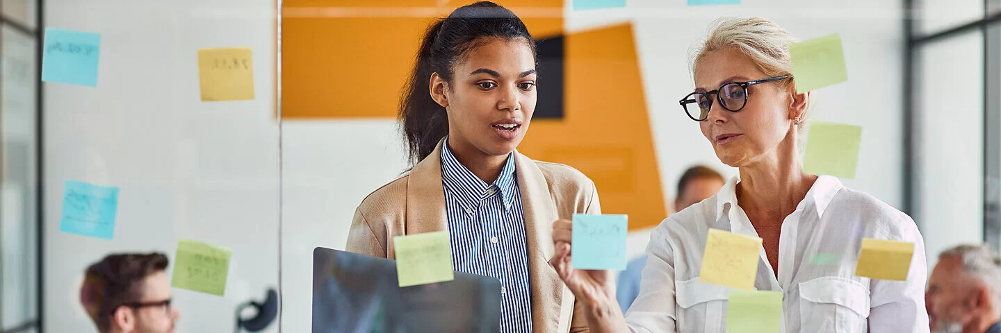 Zwei Frauen im Job schauen auf eine Glaswand mit Haftnotizen und besprechen das weitere Vorgehen.