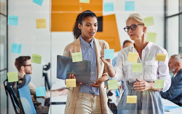 Zwei Frauen im Job schauen auf eine Glaswand mit Haftnotizen und besprechen das weitere Vorgehen.