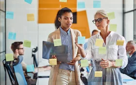 Zwei Frauen im Job schauen auf eine Glaswand mit Haftnotizen und besprechen das weitere Vorgehen.