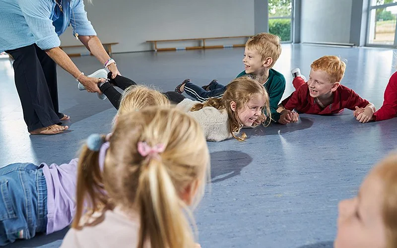 Eine Gruppe von Kindern liegt lachend und auf den Bauch gestützt auf einem blauen Turnhallenboden im Kreis. Eine Frau in hellblauem Hemd und dunkler Hose steht links und hält die Beine eines Kindes, das sich ebenfalls auf dem Boden befindet.