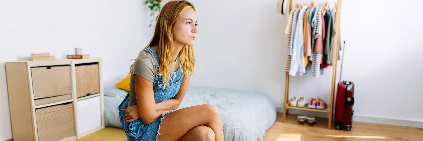 Eine junge Frau, die unter Menstruationsschmerzen leidet, sitzt in ihrem Schlafzimmer und verschränkt die Arme vor dem Bauch.  