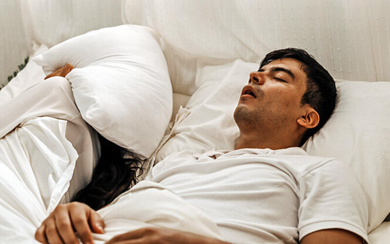 Ein Mann schnarcht im Bett und seine Partnerin hält sich ein Kissen über dem Kopf